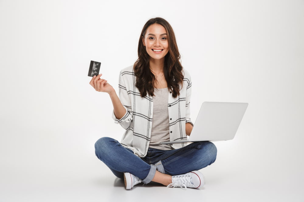 smiling brunette woman shirt sitting floor with laptop computer credit card while looking camera gray