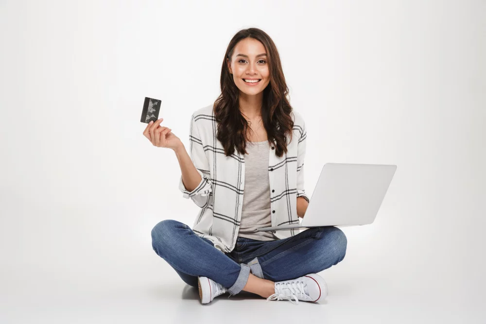 7 ações para melhorar a pontuação no score de crédito 2 smiling brunette woman shirt sitting floor with laptop computer credit card while looking camera gray